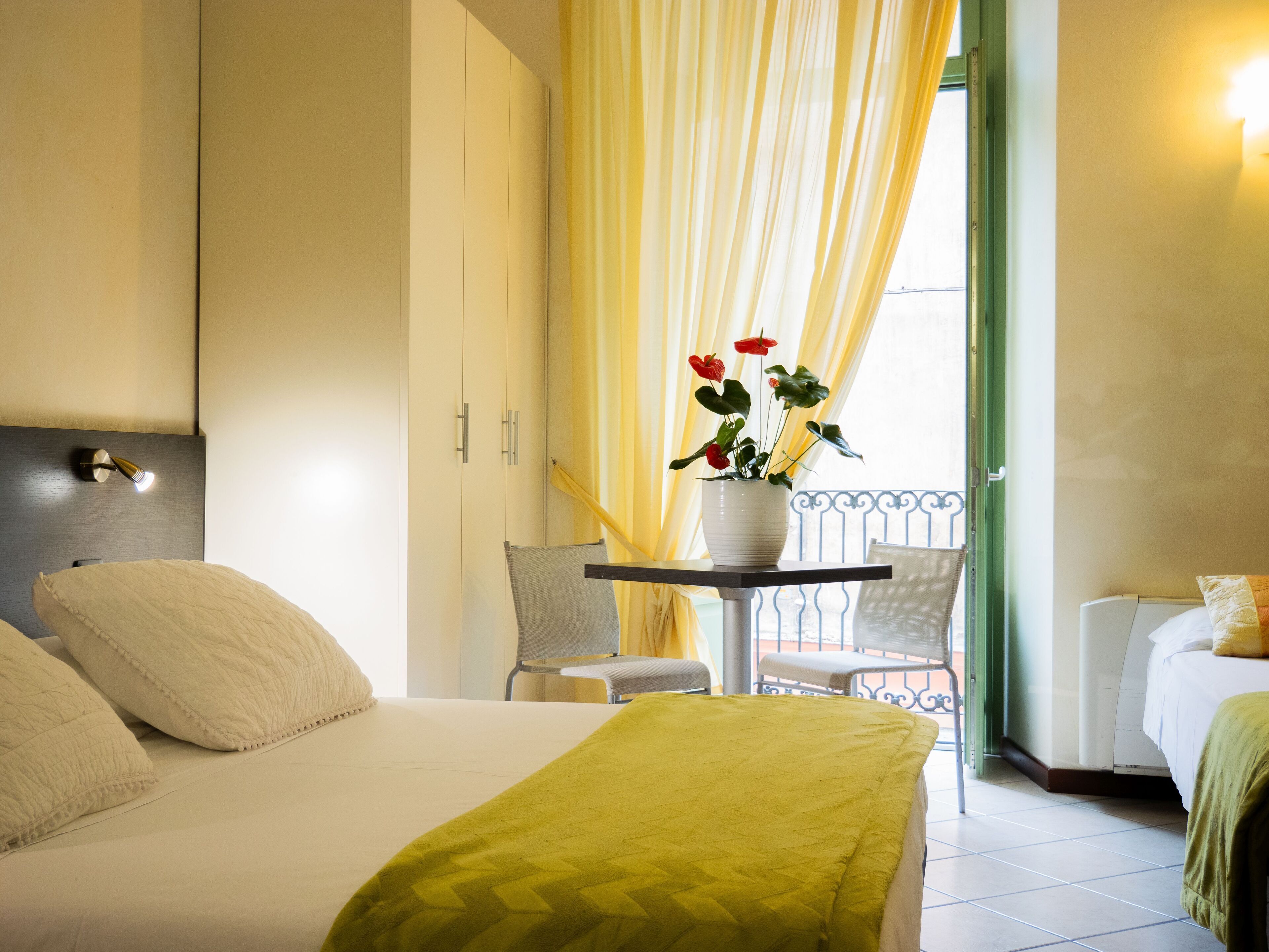 Quarto de hotel com uma cama, uma mesa pequena com uma planta em vaso, varanda com cortinas amarelas e vista para o exterior.