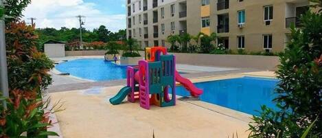 A swimming pool with a slide and play structures, surrounded by greenery and a multi-story building.
