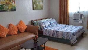 Un espacio habitable compacto con un sofá, una cama con colcha a rayas, una mesita redonda con un jarrón y una silla.