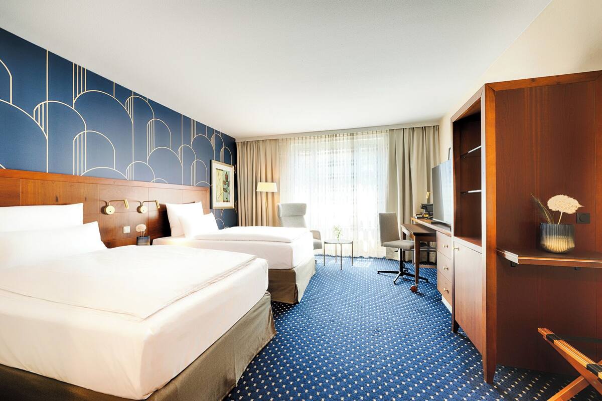 A hotel room with two beds, a desk, a chair, a lamp, and a wardrobe.
