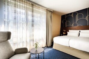 A hotel room with a large bed, a sofa, a small table with a vase, sheer curtains, and a patterned wall. - Welcome Parkhotel Bochum (Bochum)