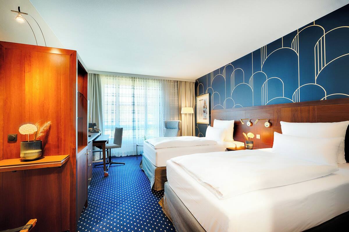 A hotel room with two beds, a desk, a chair, a wooden wardrobe, and a patterned wall.