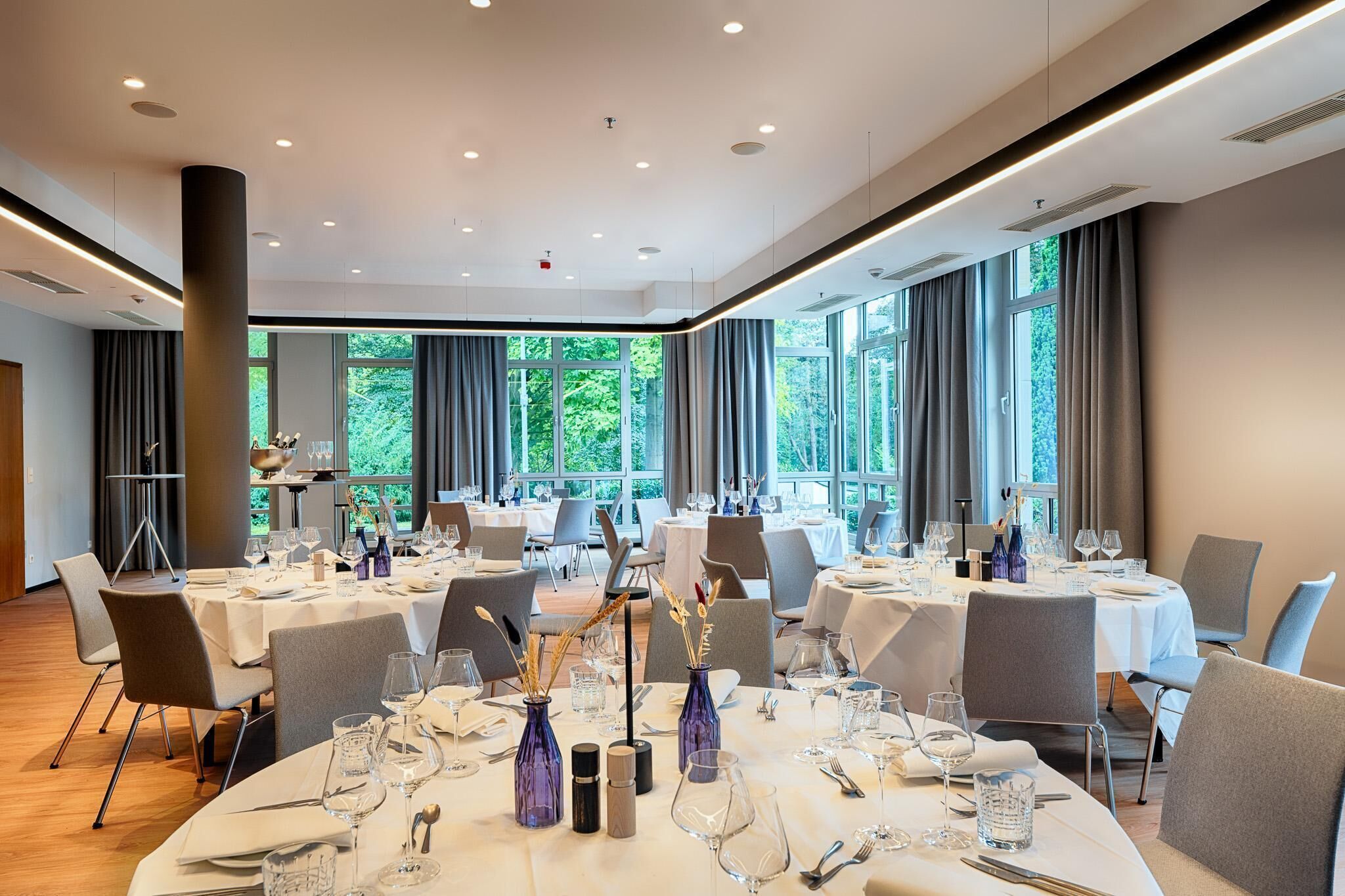 A spacious dining area with round tables set for a meal, large windows with curtains, and a modern interior design.