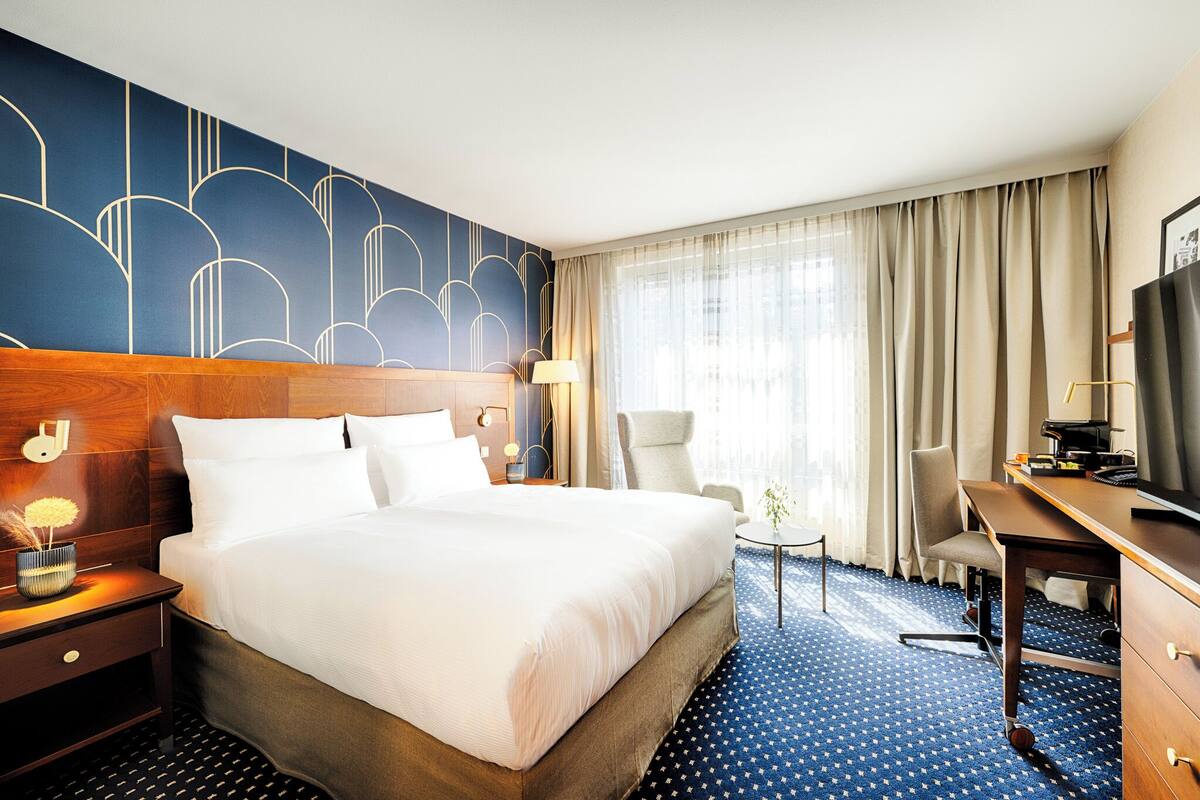A hotel room with a large bed, a desk, a chair, a TV, and a window with curtains.