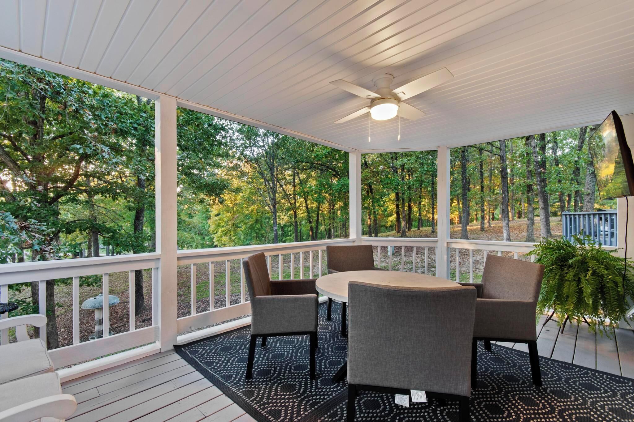 A covered outdoor area with a table and chairs, surrounded by a white railing, overlooking a wooded area.
