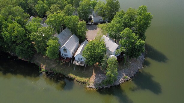 Une vue aérienne d’une maison isolée, entourée d’une forêt dense et bordée par un plan d’eau, avec une jetée qui s’avance sur l’eau.