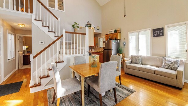 A spacious living area with a wooden staircase, a dining table with chairs, a sofa, and a kitchen in the background.