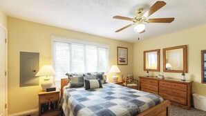 A bedroom with a wooden bed, two bedside tables with lamps, a ceiling fan, and a window with blinds. - Two Bedroom Bayside Condo (Grand Rivers)