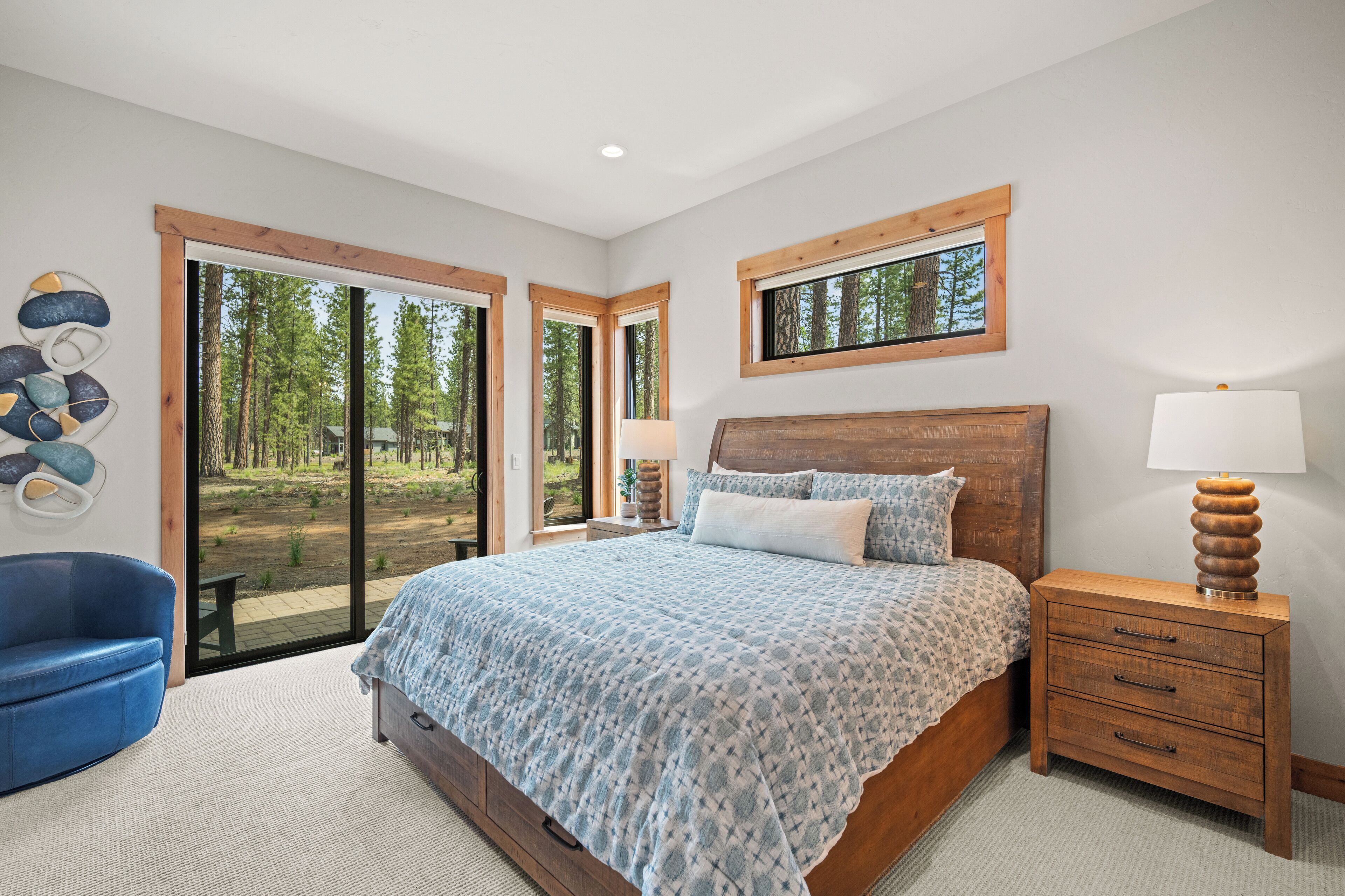 A bedroom with a large bed, wooden headboard, and a nightstand with a lamp. There is a blue chair and a window with a view of trees.