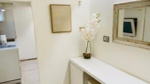 Ein Flur mit einer weißen Konsole, einem kleinen Hocker und einer Vase mit weißen Blumen. An der Wand hängt ein gerahmtes Bild und über der Konsole ein Spiegel.