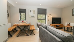 A modern living room with a wooden dining table set for four, a grey sofa, a flat-screen TV, and a floor lamp. - Brimstone (Ulverston)