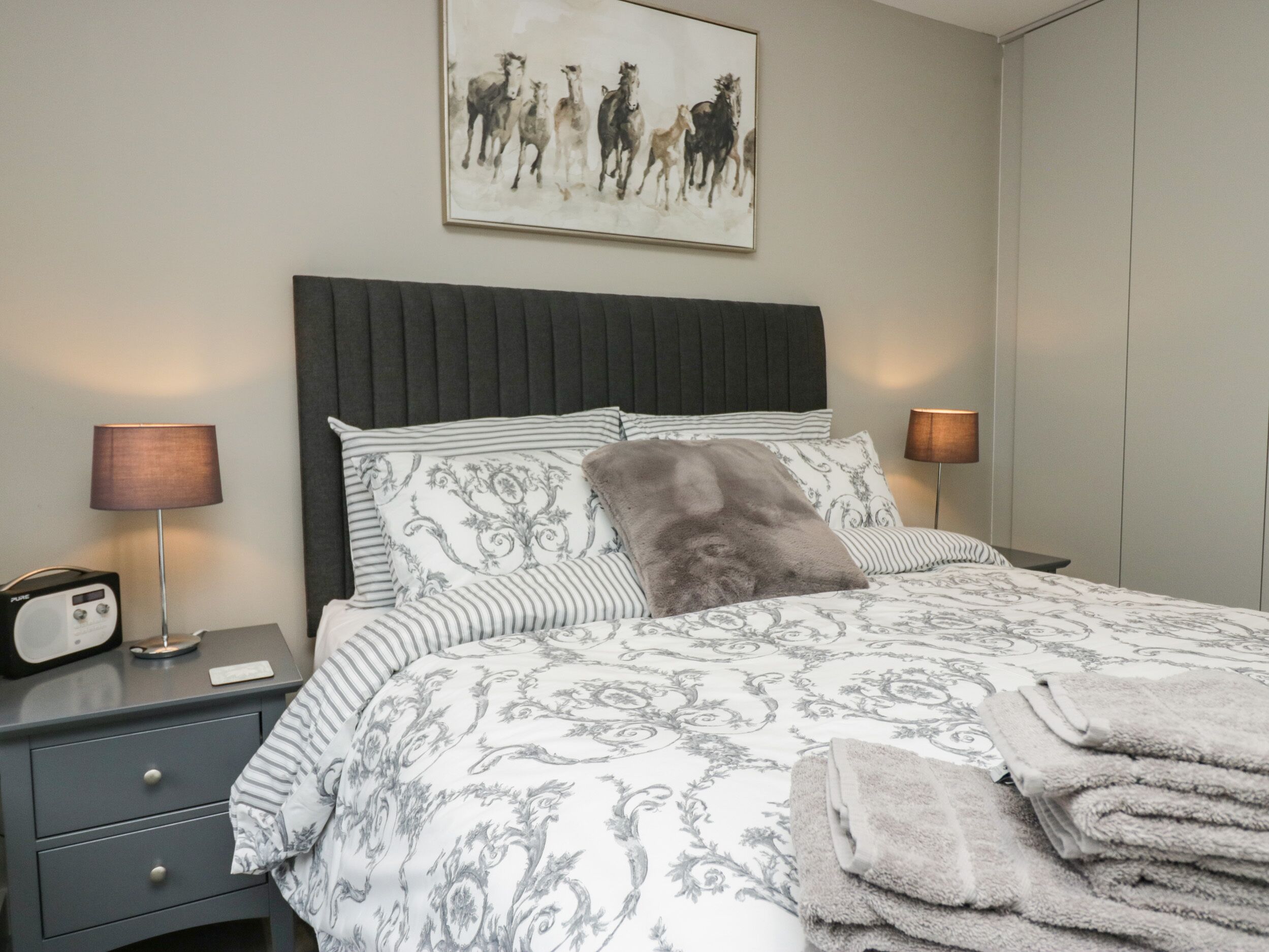 Un letto ben rifatto con una coperta a motivi, due lampade ai lati del letto, una radio e un quadro incorniciato sopra il letto.