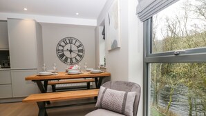 A modern kitchen with a wooden dining table set for four, a large wall clock, and a view of a river through a window. - Brimstone (Ulverston)