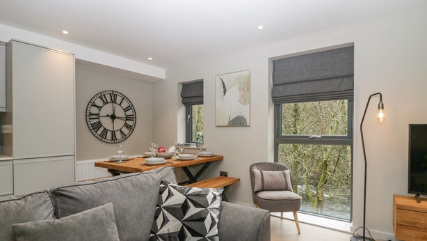 A modern living room with a grey sofa, a wooden dining table set for four, a large wall clock, and a window with a view of trees. - Brimstone (Ulverston)