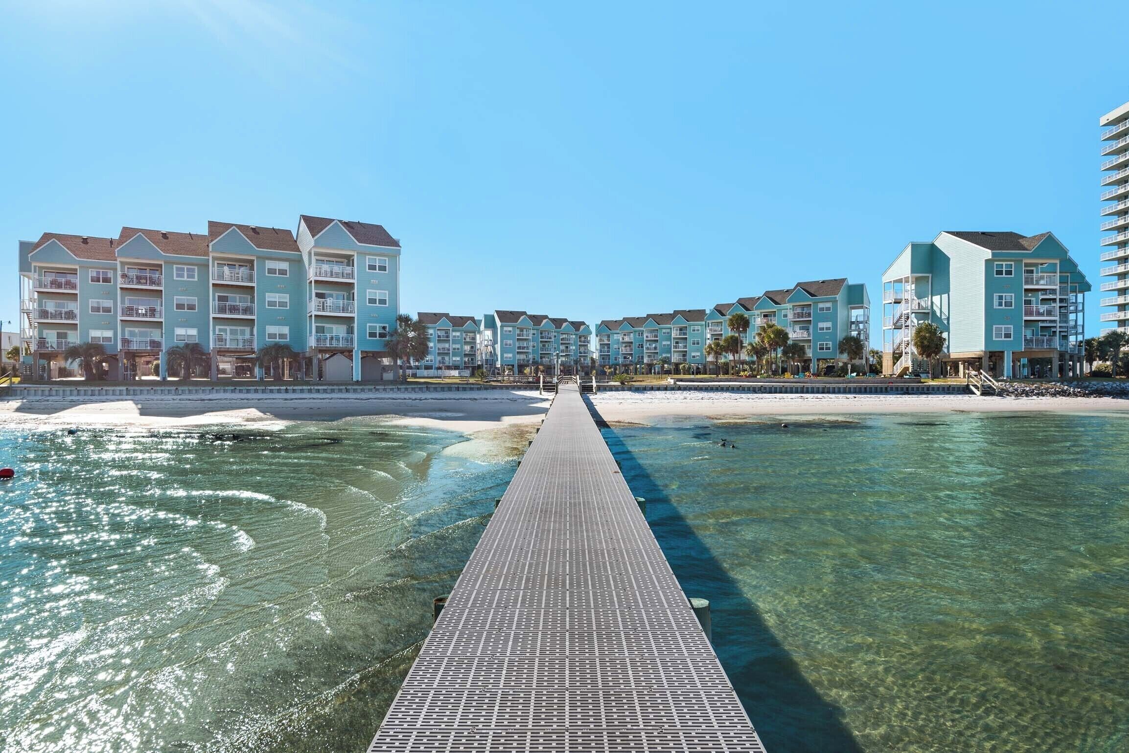 Un resort con un paseo elevado sobre aguas claras que conduce a una playa, rodeado de edificios de varios pisos con balcones.