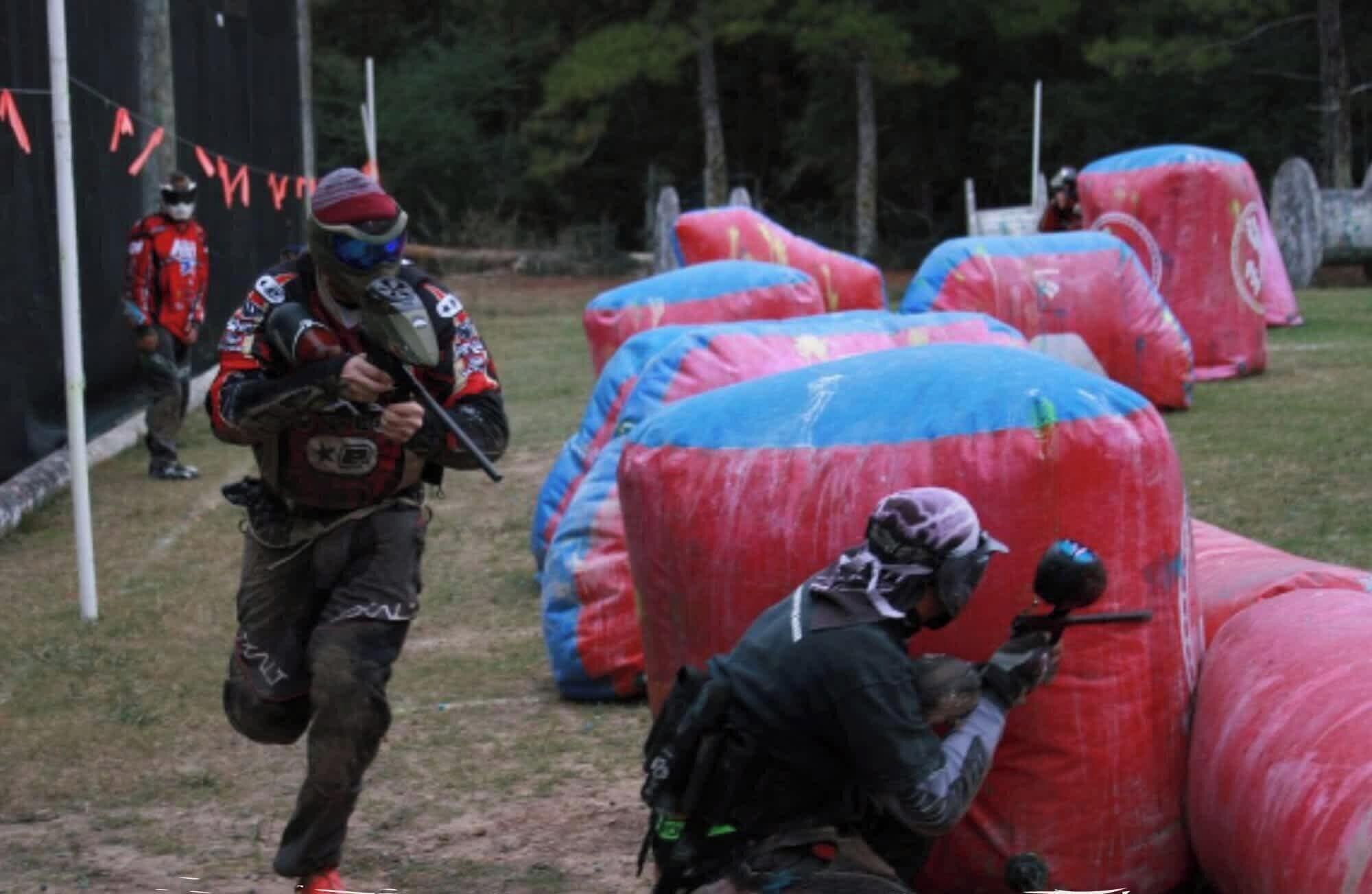 A group of people engaged in a paintball game, with inflatable barriers and trees in the background.