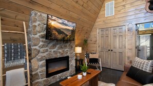 Un interior de cabaña acogedor con chimenea de piedra, televisor montado, un banco y una puerta.