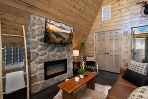L’intérieur chaleureux d’un chalet avec une cheminée en pierre, un téléviseur fixé au mur et un plafond en bois.