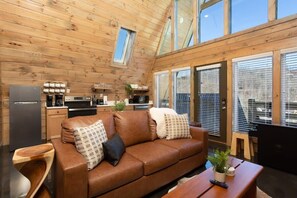 L’intérieur chaleureux d’un chalet avec un plafond en bois, un coin cuisine, un canapé en cuir marron et un espace repas.