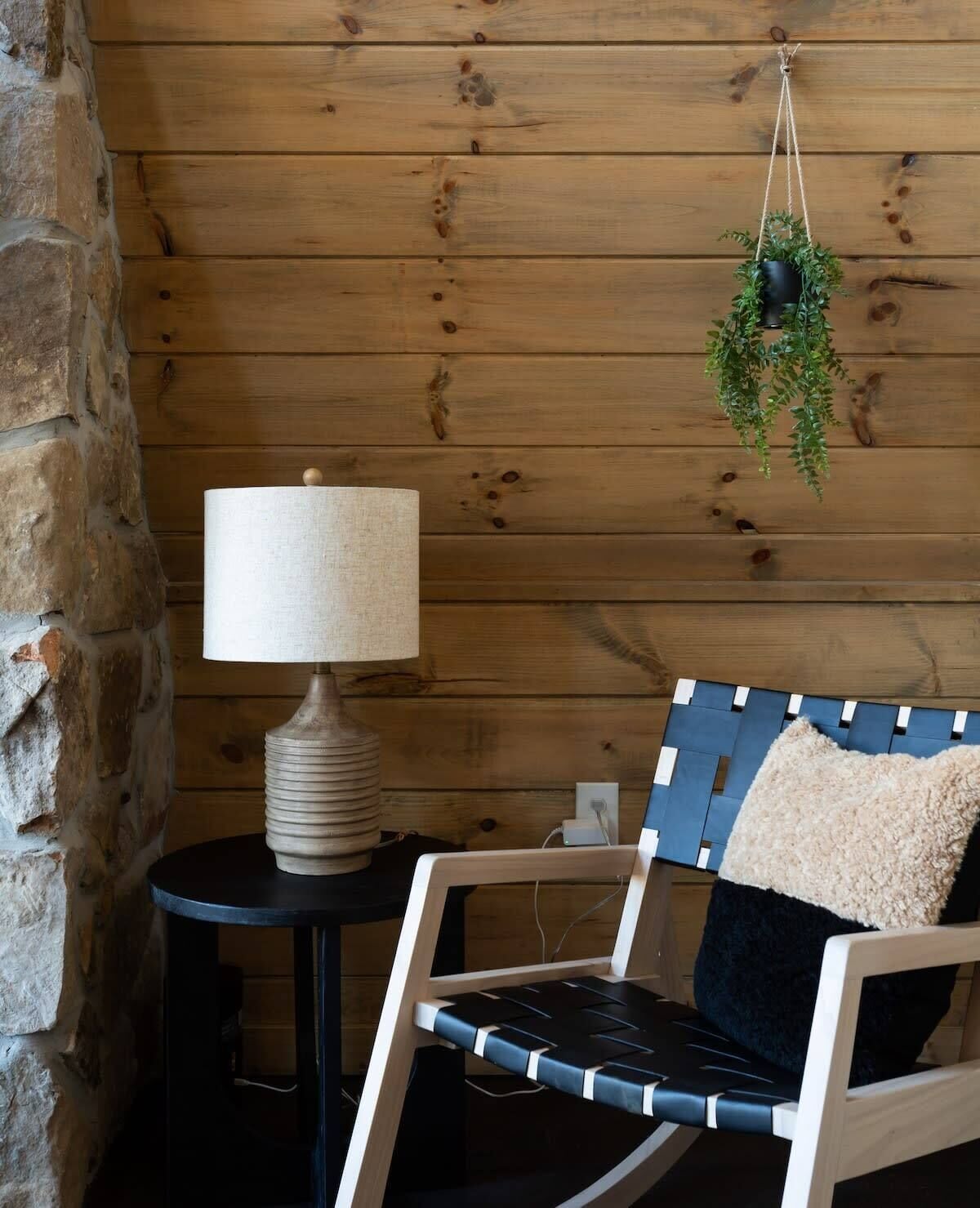 Una pared de madera, una lámpara, una silla con cojín y una planta colgante.