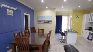 A dining area with a large wooden table and chairs, a kitchen with white cabinets, and a living area with a black sofa. - The Shack at Stansbury - Short walk to the beach! Off peak special! (Stansbury)