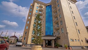 A multi-story building with a prominent blue glass facade, a central entrance with a canopy, and a parking area with cars. - Watercress Hotels and Signature (Lagos)