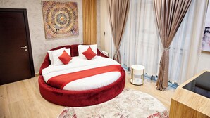 A modern bedroom with a circular bed, a large abstract painting, and a view of the city through sheer curtains. - Watercress Hotels and Signature (Lagos)