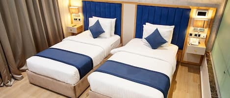 A hotel room with two beds, each with a blue headboard, white bedding, and a blue accent pillow. There are bedside tables with lamps, a painting on the wall, and a wooden floor.