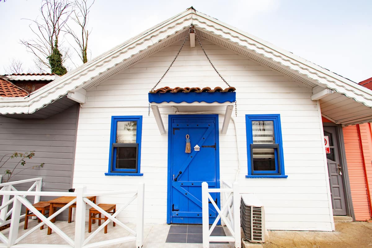 A white building with a blue door and windows, a small porch with wooden furniture, and a tiled roof.