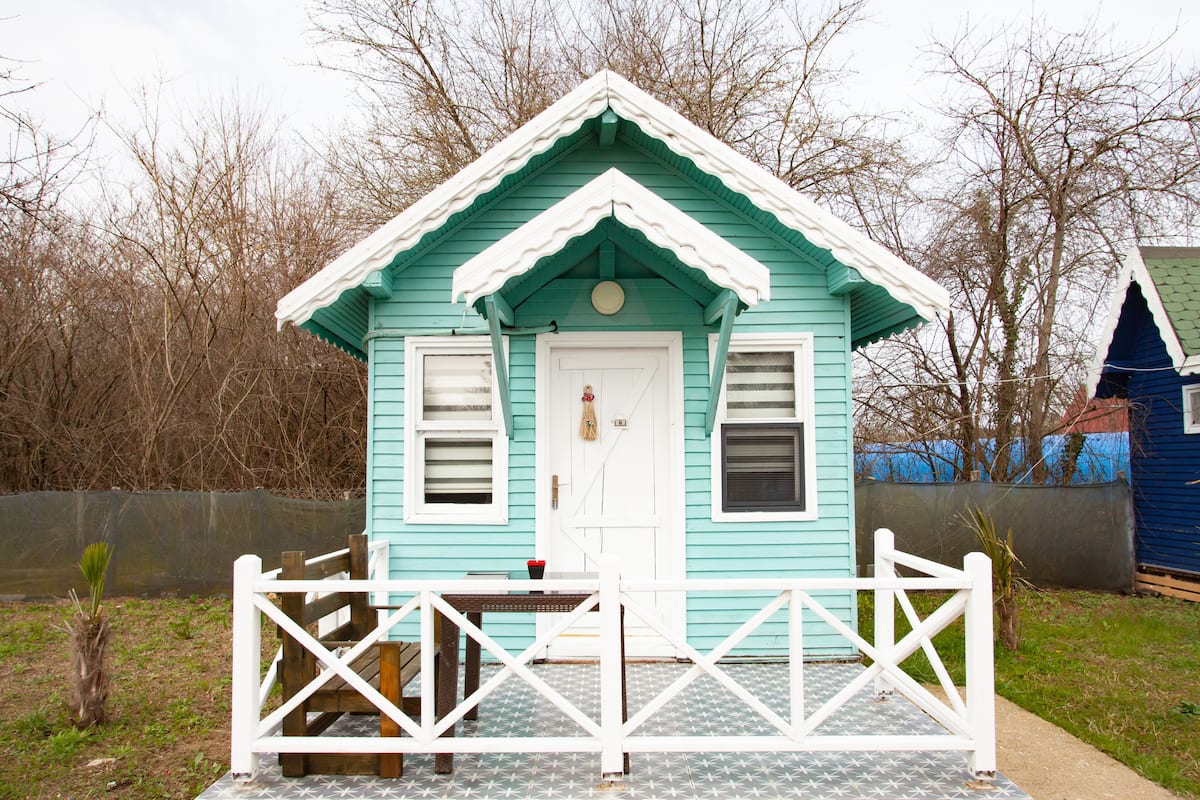 A small, light blue house with a white door and windows, a porch with a white railing, and a small table with a chair.
