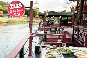 Un área de comedor junto al río con mesas y sillas, un espacio de estar cubierto y un cartel que dice "TIAS Shelae Otel".