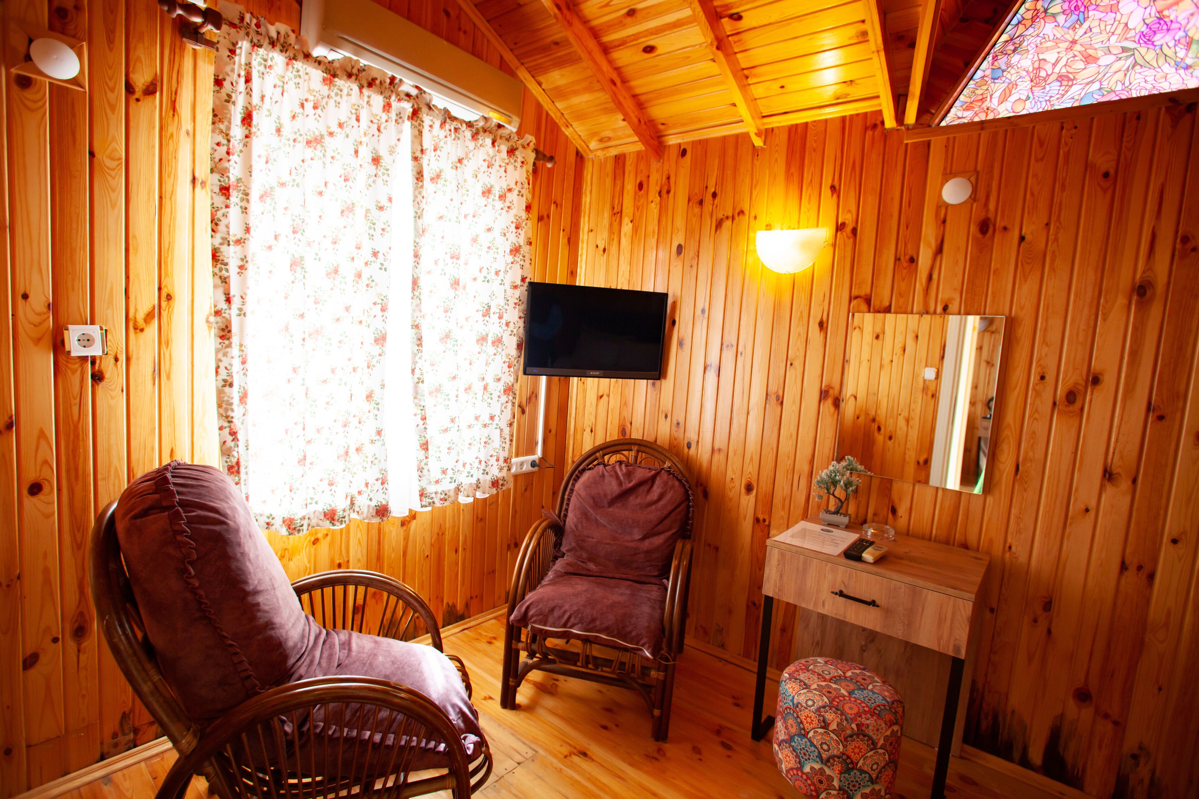 A wooden room with two chairs, a small table, a television, and a window with floral curtains.