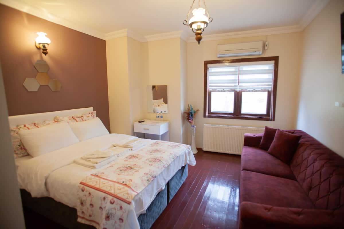 A hotel room with a bed, a red sofa, a bedside table, a mirror, and a window with blinds.