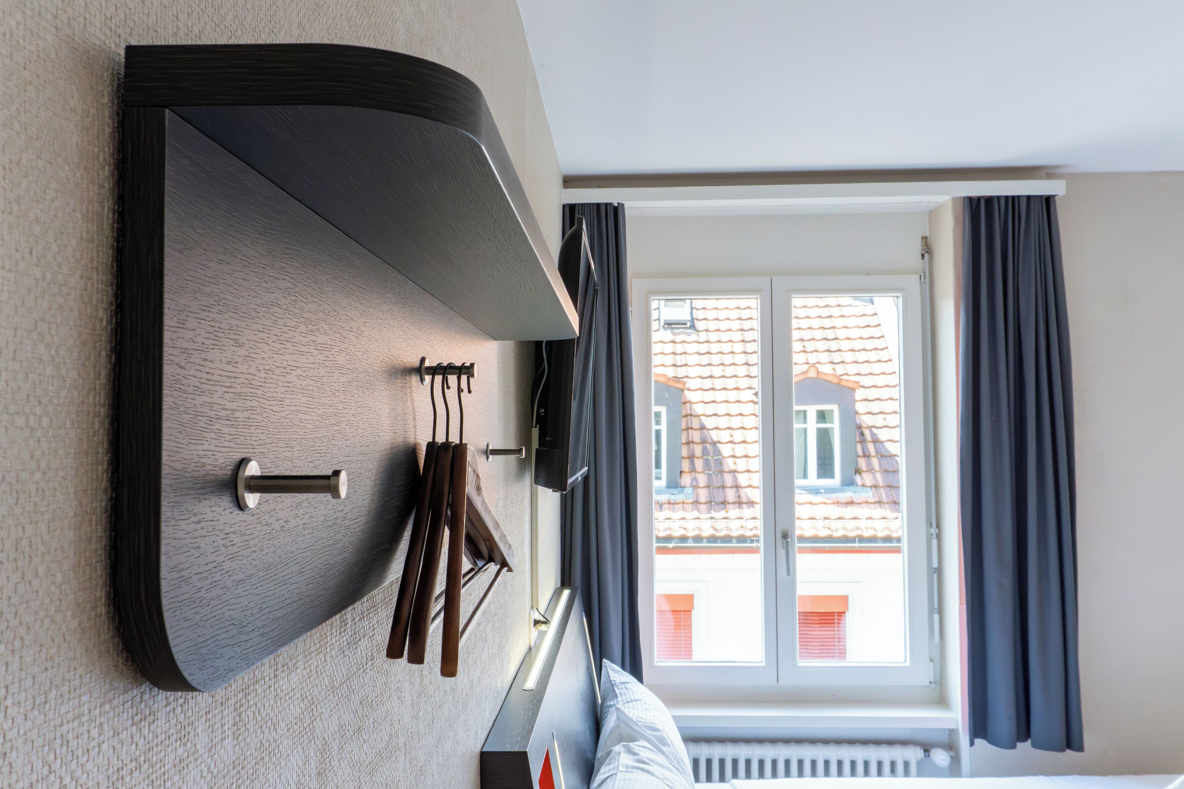 A hotel room with a built-in shelf, a window with curtains, and a view of buildings outside.