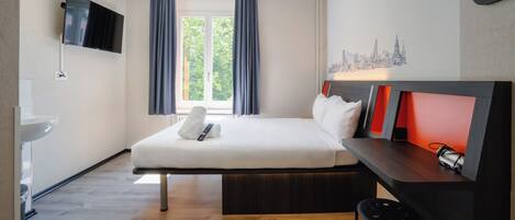 Una habitación de hotel moderna con una cama, un escritorio, un televisor, un ventanal con cortinas y un cuadro de arte con un paisaje urbano.