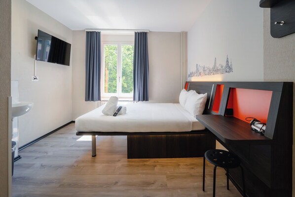 Una habitación de hotel moderna con una cama, un escritorio, un televisor, un ventanal con cortinas y un cuadro de arte con un paisaje urbano.