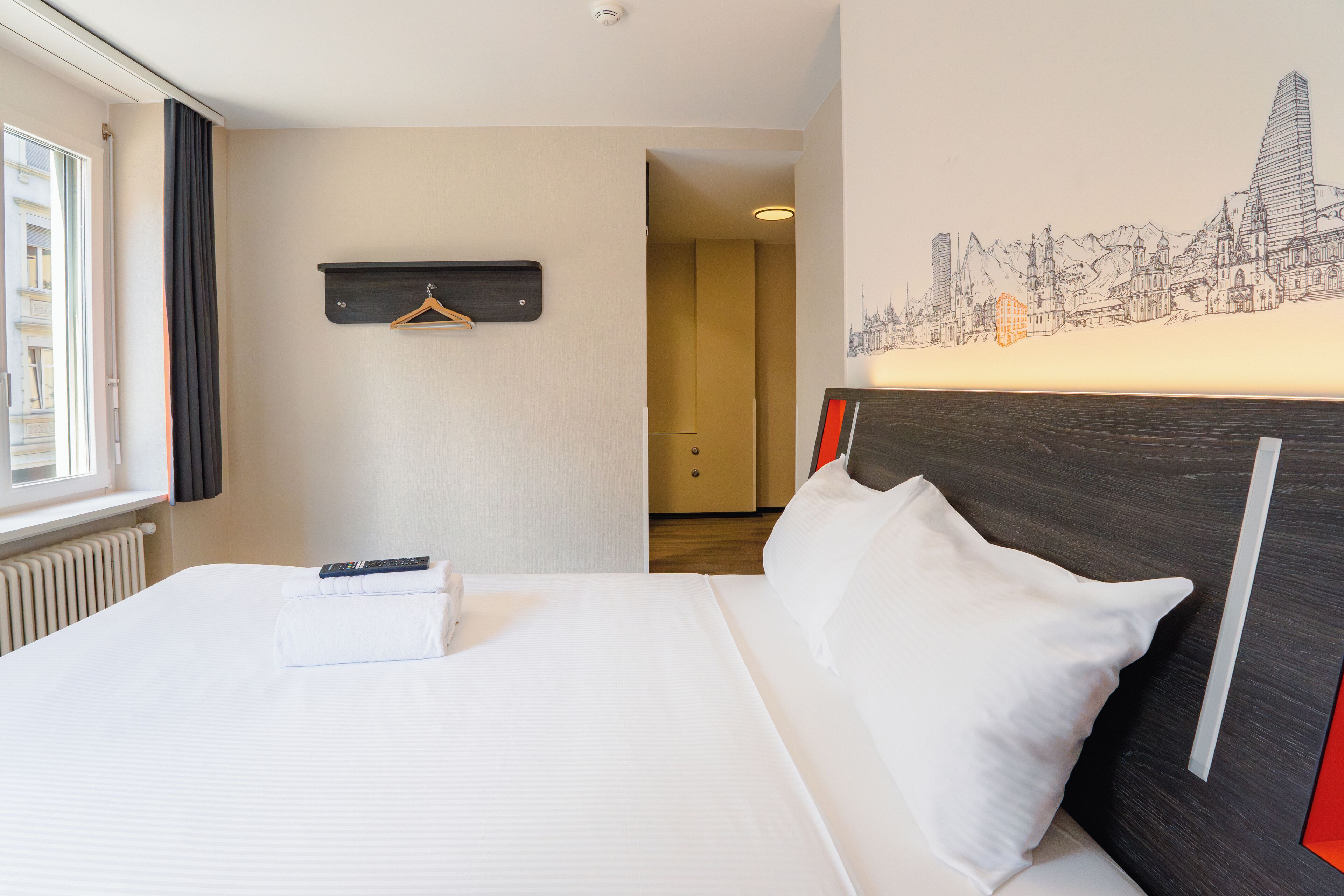 A hotel room with a neatly made bed, a remote control, a hanging coat rack, and a cityscape mural on the wall.