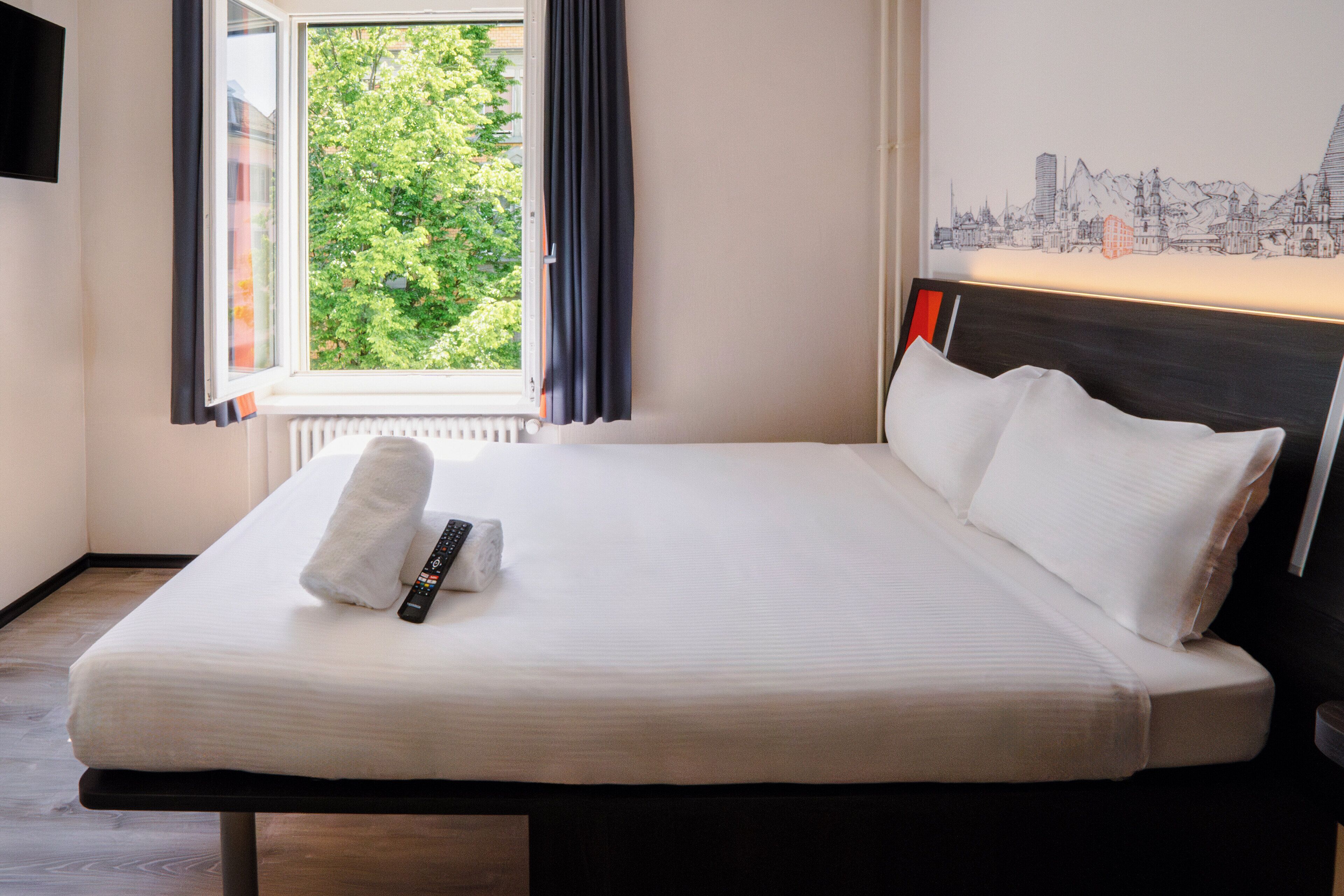 A neatly made bed with white linens and a remote control, a window with curtains, and a cityscape artwork on the wall.