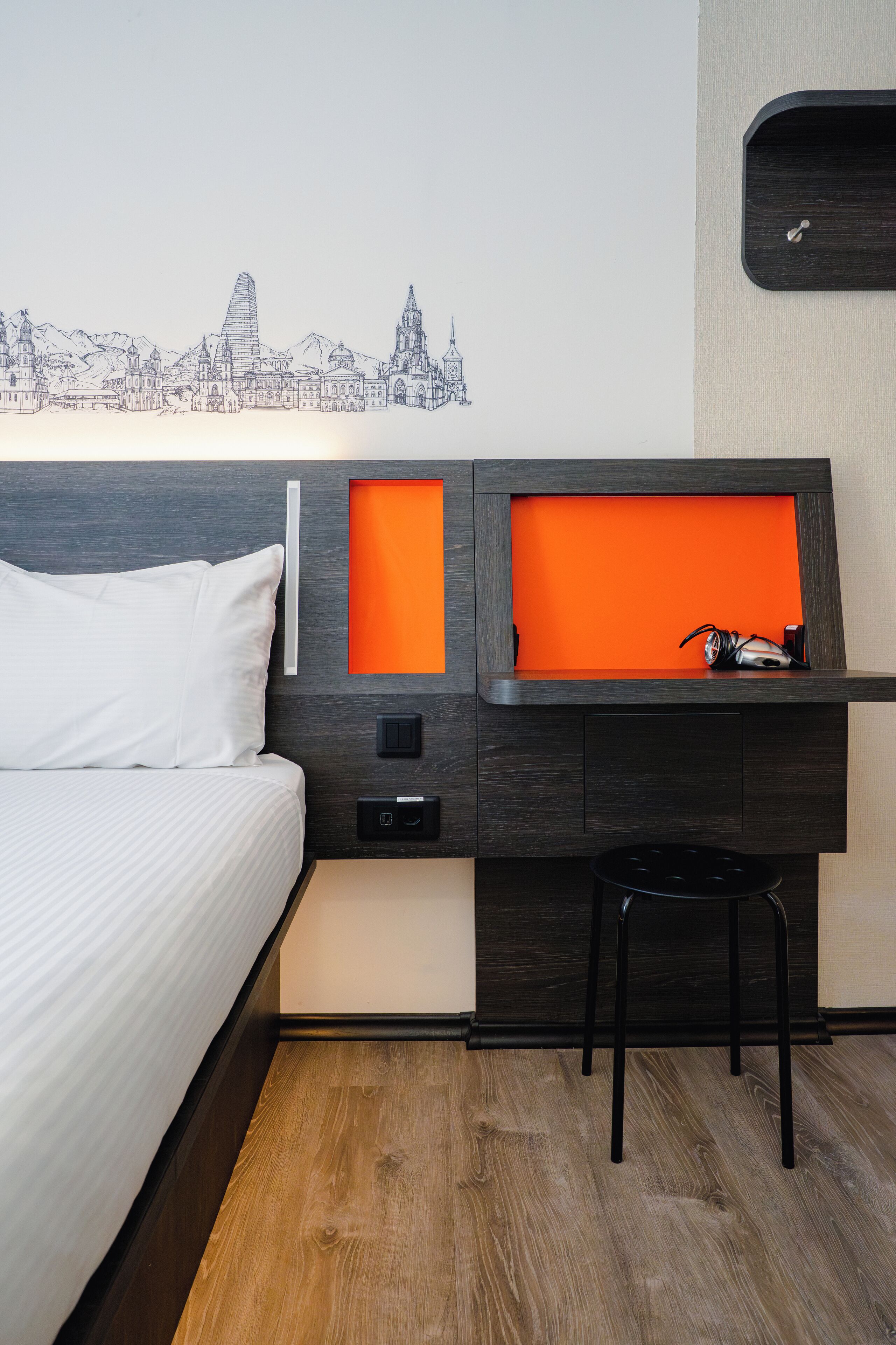 Une chambre d’hôtel avec un lit, un bureau orné d’une fresque représentant un paysage urbain, une chaise et une petite étagère orange.