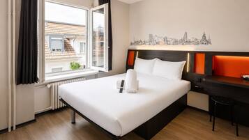 Camera d'hotel con un letto, una scrivania, una sedia, una finestra con tende e un quadro raffigurante un panorama urbano.