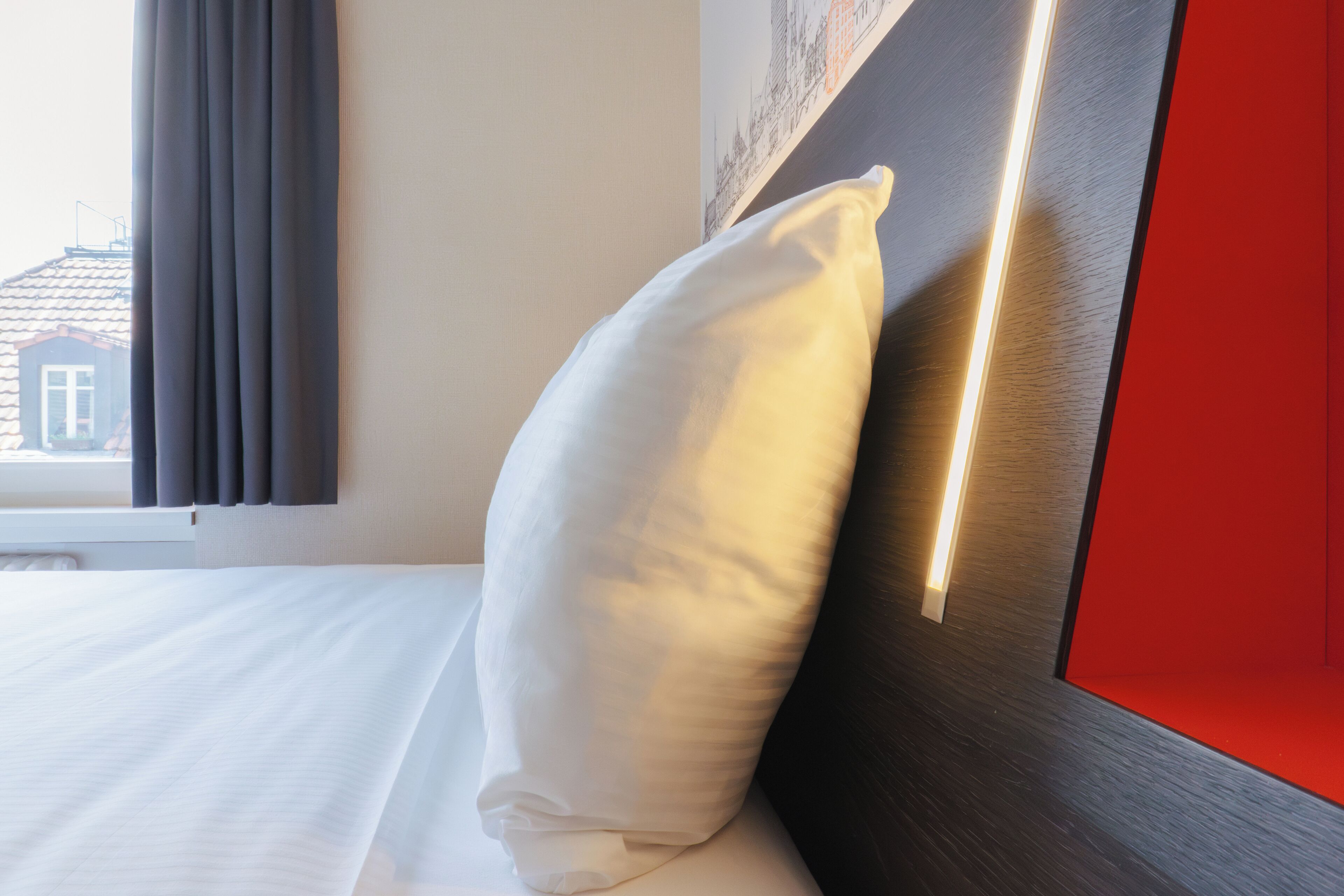 Una cama bien tendida con un almohadón blanco, un cabecero oscuro con una tira de luz, y una ventana con cortinas.