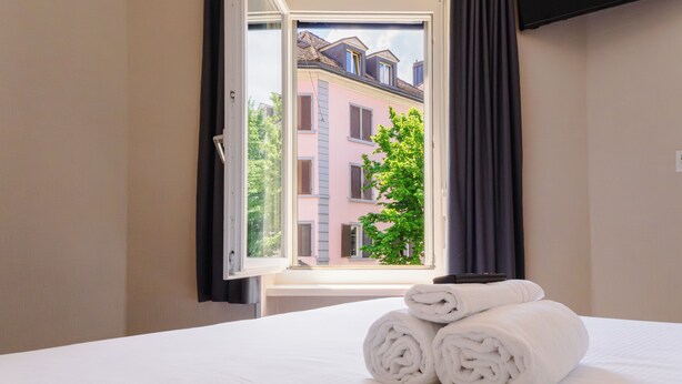 A hotel room with a bed, a window with curtains, and a view of a building outside.
