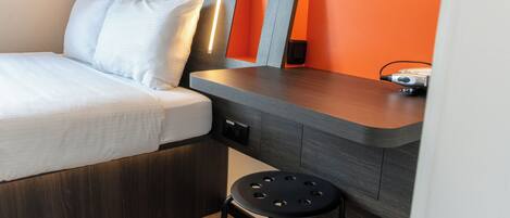 A modern hotel room with a bed, a small stool, and a desk with a lamp.