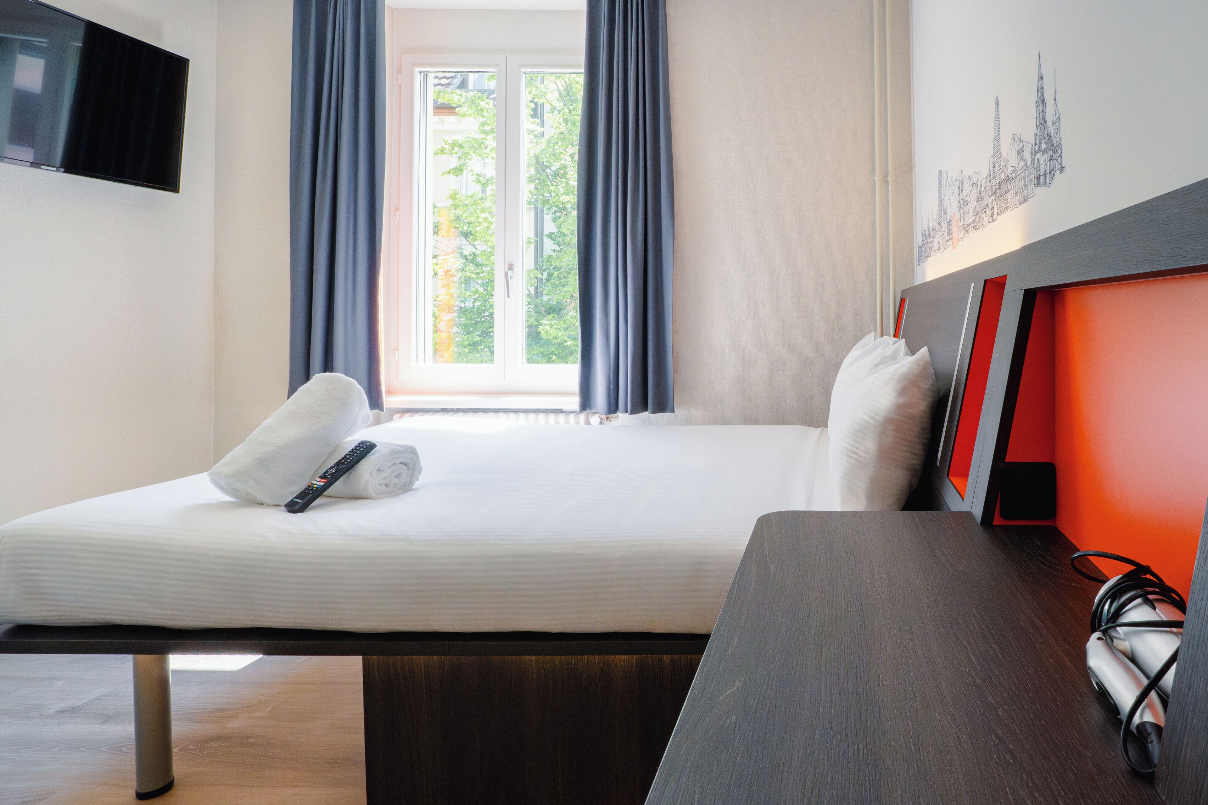 A hotel room with a bed, a TV mounted on the wall, a window with curtains, and a cityscape artwork on the wall.