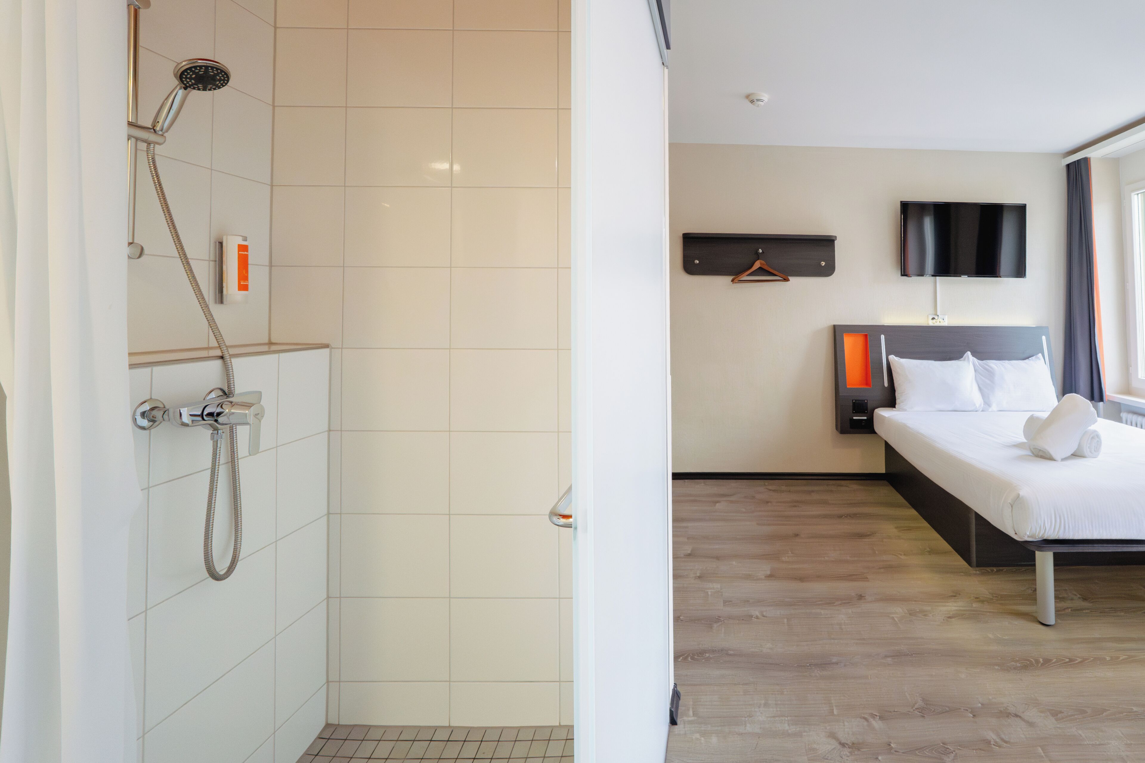 Una habitación de hotel con ducha, una cama con sábanas blancas, un televisor montado y piso de madera.