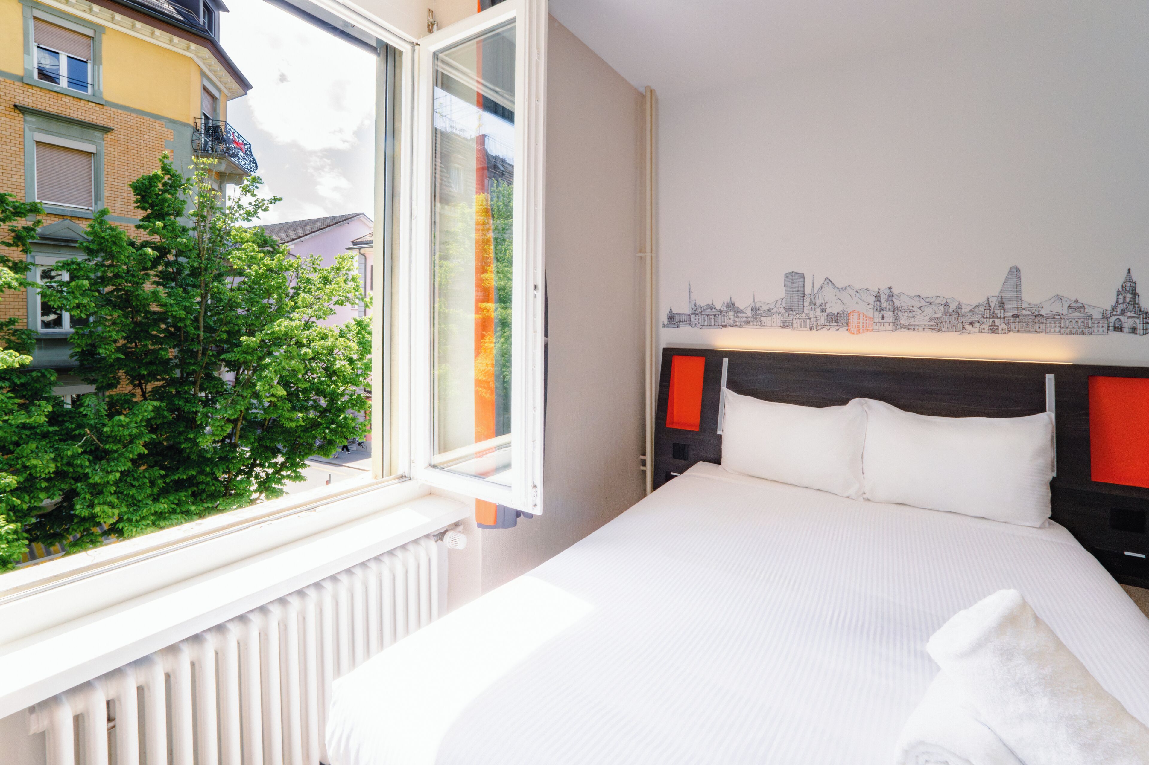 A hotel room with a bed, a window with a view of a tree and buildings, and a cityscape mural on the wall.