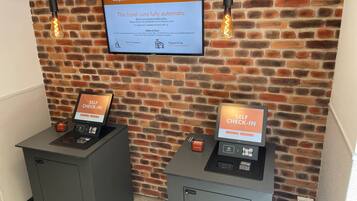 A self-check-in kiosk and a television displaying hotel information against a brick wall backdrop.