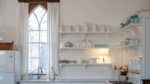 Een keuken met witte betegelde muren, een raam met witte gordijnen en planken met keukengerei.