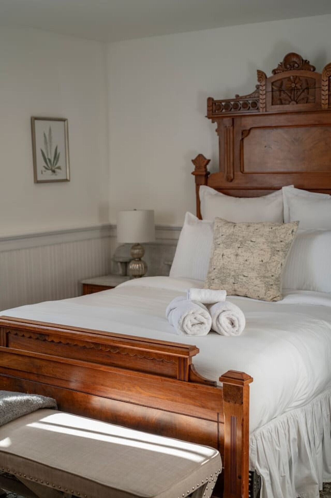 A neatly made bed with white linens and rolled towels, a wooden headboard with intricate carvings, and a small bedside table with a lamp.