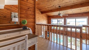 A wooden interior with a desk, chair, and a view of the beach through large windows. - Cavallo Dunes - 4x4 Carova Beach, Oceanfront, Ocean View, Hot Tub, Pets OK, Year Round Flex Stays (4x4 - Carova Beach)